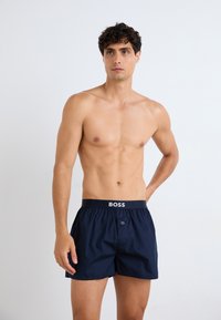 Marineblå boxershorts laget av lett stoff; har elastisk midjebånd med "BOSS"-logo og en knapp i front for lukking.