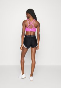 Αθλητικό σουτιέν σε φωτεινό ροζ χρώμα με διασταυρωμένες τιράντες, συνδυασμένο με μαύρες αθλητικές σορτς που διαθέτουν άσπρη ρίγα και λογότυπο στη ζώνη.