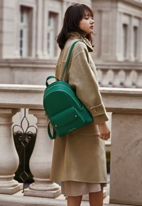 Green leather backpack with a rounded shape, featuring a front pocket and gold hardware accents, worn on a beige coat.
