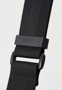Sangle en nylon noir présentant un motif texturé et une boucle en plastique noir mat avec le logo "BOSS" en relief. Ajustable en longueur.