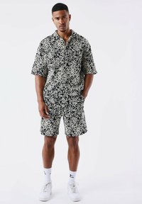 Camisa negra de manga corta y pantalones cortos a juego con patrones florales blancos, con un corte holgado y dos bolsillos laterales. Zapatillas blancas puestas.