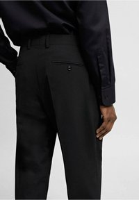 Czarne spodnie o gładkiej fakturze, z zapinanym na guzik tylnym kieszonką i eleganckim, dopasowanym krojem. Noszone z czarną koszulą z długim rękawem.
