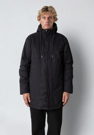 Blond muž v černém zimním kabátě s kapucí, zapínání na přední zip a stahovacími šňůrkami, stojící před jednobarevným světlým pozadím.