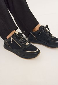 Sneaker neri con accenti dorati, dotati di una tomaia texturizzata, cerniere decorative e una suola in gomma. I lacci sono intrecciati con filo dorato.