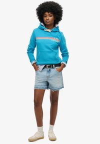 Turquoise hoodie met een voorkleurzak, witte trekkoord en een regenboogpatroon, gecombineerd met lichtgekleurde denim short en crème sneakers.