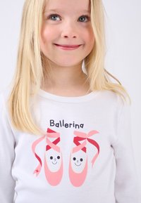 Camicia bianca a maniche lunghe con una grafica di due scarpette da balletto rosa con nastri rossi e volti sorridenti, con la scritta "Ballerina" sopra.
