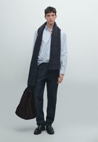 Camicia a righe blu chiaro, sciarpa di lana scura con frange, jeans scuri, scarpe di pelle nere e tiene in mano una borsa marrone scuro. Composizione semplice.