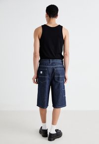 Denim shorts in dark blue with a relaxed fit, four pockets, contrasting white stitching, paired with a black tank top and black shoes.