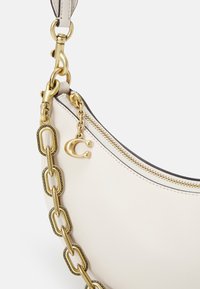 Coin de sac à main en cuir blanc avec bandoulière chaîne dorée, fermeture éclair dorée et pendentif en lettre "C" dorée suspendue.