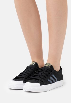 Schwarze Leinen-Turnschuhe mit weißen Gummispitzen und -sohlen, die blaue Streifen an der Seite und schwarze Schnürsenkel haben. Goldene Logo-Akzente auf der Zunge.