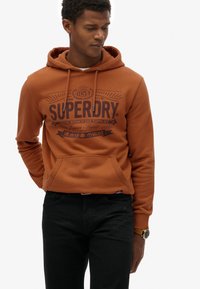 Oranje hoodie van fleece stof met een zak aan de voorkant en een geprinte logo in donkerbruin. Gekocht met zwarte jeans.