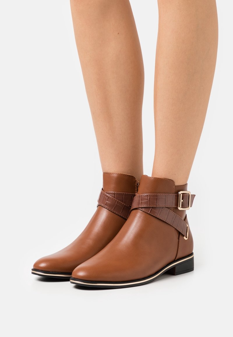 Bottes courtes marron avec bouts pointus et sangles texturées enroulées, attachées par des boucles dorées, portées sur des jambes nues sur un fond blanc.
