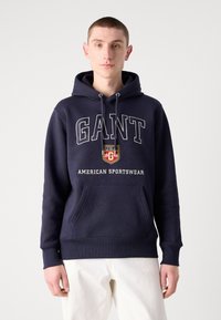 Sudadera de color azul marino hecha de mezcla de algodón, con un bolsillo frontal tipo canguro, capucha con cordón y letras blancas "GANT AMERICAN SPORTSWEAR".