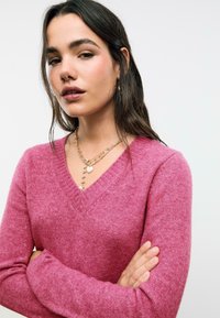 Žena s dlouhými tmavými vlasy, oblečená v růžovém svetr s výstřihem do V a vrstvenými zlatými náhrdelníky ve tvaru srdce, s crossed arms na jednoduchém bílém pozadí.