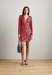 Robe mini en sequins rouges avec des manches longues et un design croisé. Le mannequin porte un petit sac argenté et des sandales à talons noires. Fond en parquet.
