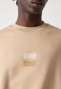 Gros plan sur une personne portant un sweat-shirt beige à col rond avec un logo carré bicolore "HUGO" sur la poitrine.