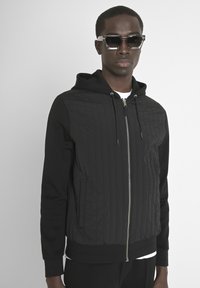 Giacca nera con cappuccio, con texture trapuntata sul davanti, tessuto liscio sulle maniche e zip frontale. Indossata sopra una camicia bianca.