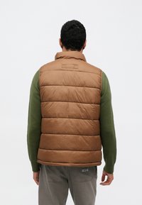 Barna steppelt, vízszintes mintázatú mellény, magas nyakkal és sima textúrával. Zöld hosszú ujjú ing fölött viselve.