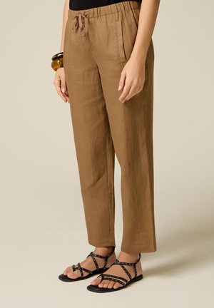Pantaloni marroni a gamba dritta con vita con coulisse indossati con sandali bassi neri con borchie su una persona in piedi.