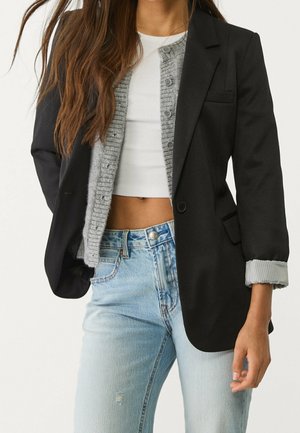 Vrouw die lichtblauwe jeans, een wit cropped top, een grijze cardigan met knopen en een zwarte blazer met opgerolde mouwen draagt.