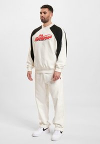 Hvid sweatshirt med sorte raglanærmer, prydet med rød grafisk tekst. Kombinationen kompletteres med matchende hvide sweatpants og hvide sneakers med sorte detaljer.