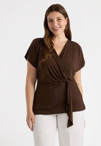 Top marrón con mangas cortas, detalle de lazo en la cintura, hecho de una tela suave. Combinado con pantalones blancos, de textura lisa y corte relajado.