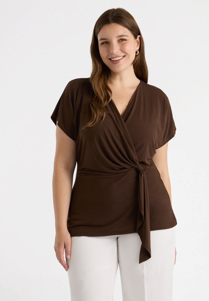 Top marrón con mangas cortas, detalle de lazo en la cintura, hecho de una tela suave. Combinado con pantalones blancos, de textura lisa y corte relajado.