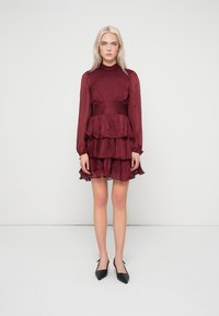 Κουμπαρίσια, φόρεμα με μακριά μανίκια και ψηλό λαιμό, με ριχτό φούστα σε στρώσεις και βελονιά στην μέση. Φορεμένο με μαύρες στιλέτο γόβες.