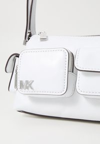 Biela kožená crossbody taška so strieborným kováním, obsahujúca hlavnú priehradku na zips a predné vrecko s prešívanými detailmi a logom.