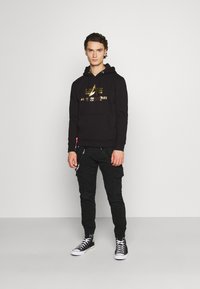 Svart hettegenser med gulltrykket logo, kombinert med svarte cargo-bukser og svarte høye sneakers med hvite såler. Enkel, slank design.