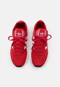 Chaussures de sport rouges en maille, dotées de trois bandes blanches, d'une pointe arrondie, de lacets texturés et d'une semelle blanche contrastante.