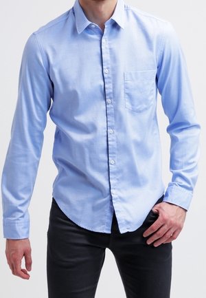 Man wearing a light blue long-sleeve button-up shirt with a front pocket and black pants, standing against a plain background.