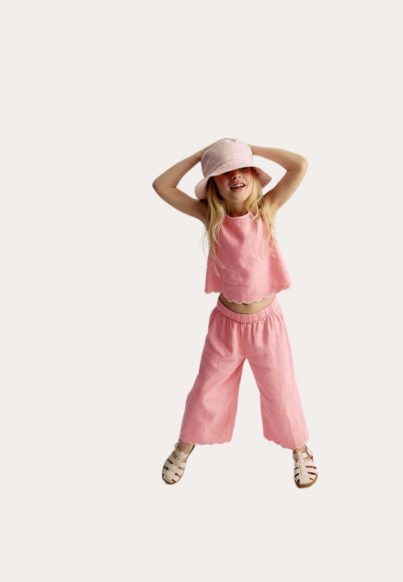 Crop top en lin rose avec un ourlet festonné, pantalon large assorti et un chapeau seau rose. Des sandales beiges à lanières complètent la tenue.