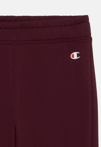 Burgunderfargede joggebukser med teksturert stoff, elastisk midjebånd og en liten brodert logo i rødt og hvitt på venstre side.