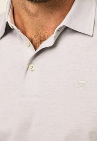 Polo léger gris fabriqué en tissu texturé, avec un col, trois boutons et un petit logo brodé sur la poitrine gauche.