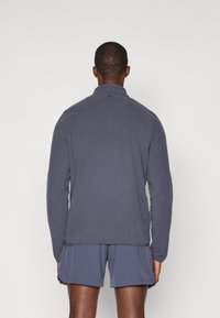 Vīriešu pelēks flīsa žakets ar augstu apkakli un garām piedurknēm. Tam ir vienkārša tekstūra un nedaudz brīvs piegriezums, savienots ar atbilstošām šortiem.