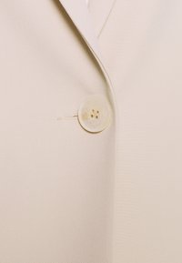 Veste beige en tissu texturé, fermeture à un bouton avec quatre trous, et col à revers ajusté pour un look cintré.