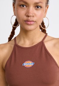 Brun halterneck-top laget av glatt stoff, med en sirkulær blå og gul "Dickies"-logo på brystet. Enkelt design med tynne stropper.