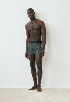 Atletisk ung mand, stående barfodet mod en hvid væg, iført mønstrede sort-hvide boxershorts.