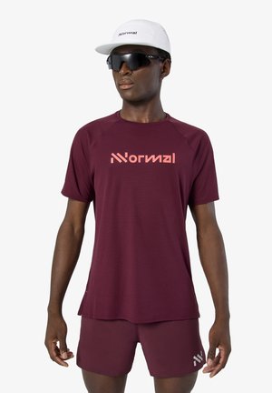 Hombre con camiseta y pantalones cortos deportivos color granate con el logo "Normal", gorra blanca y gafas de sol deportivas negras, posando frente a un fondo blanco.