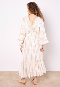 Fließendes cremefarbenes Maxikleid mit goldenen Paisleymustern, V-Rücken-Design und einer Kordelzug-Waist. Texturiertes Material mit weiten Ärmeln.