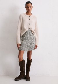 Beige gebreide cardigan met zwarte knopen, grijze tweed rok en bruine suède laarzen tot de knie met een puntige bovenrand.