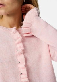 Pink cardigan made of soft material, featuring a ruffled edge along the button line and silver buttons at the front. Long sleeves with ribbed cuffs.