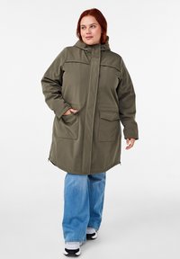 Olijfgroene buitenjas met een capuchon, voorzien van voorzakken en een rechte snit. Gepaard met lichtblauwe wijde jeans en zwarte sneakers.