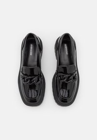 Musta patentoitu nahkakengät, joissa on paksu pohja, koristeellinen ketjuvipa sekä klassinen loafer-malli. Sujuva, kiiltävä pinta.
