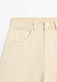 Beiger, hoch taillierter Jeansrock mit vorderer Knopfleiste, Gürtelschlaufen und aufgestickten Taschen vorne vor weißem Hintergrund.