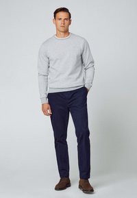 Grå crewneck sweatshirt med vita accenter, kombinerad med mörkblå byxor och bruna skor, stående mot en neutral bakgrund.