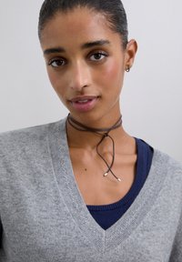 Mladá žena s vlasy stáhnutými do culíku, oblečená v šedém svetr s V-výstřihem přes námořnickou halenku a tenkým hnědým chokerem.