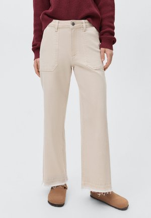 Beige wijde broek met voorzakken, rafelige zoom en knoopsluiting; gecombineerd met een kastanjebruine gebreide trui en bruine schoenen.