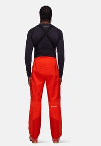 Pantalons de ski rouges en tissu durable, avec un soutien noir en croix au dos, une taille ajustable et des jambes inférieures renforcées pour une protection supplémentaire.